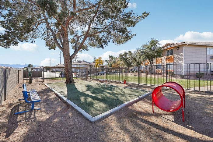 Charming community play area with vibrant red play tunnel, lush green lawn, and towering tree providing shade in a bright and sunny apartment complex.