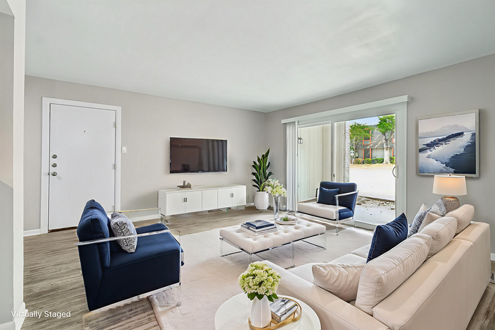 Bright and modern living room with elegant decor, featuring a sleek white sofa, plush navy chairs, and a glass-top coffee table. Large sliding doors open to a lush outdoor view, enhancing the room's welcoming atmosphere.