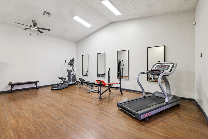 Bright and modern fitness room with sleek wooden flooring, featuring a treadmill, elliptical machine, weight bench, and wall mirrors for an enhanced workout experience.