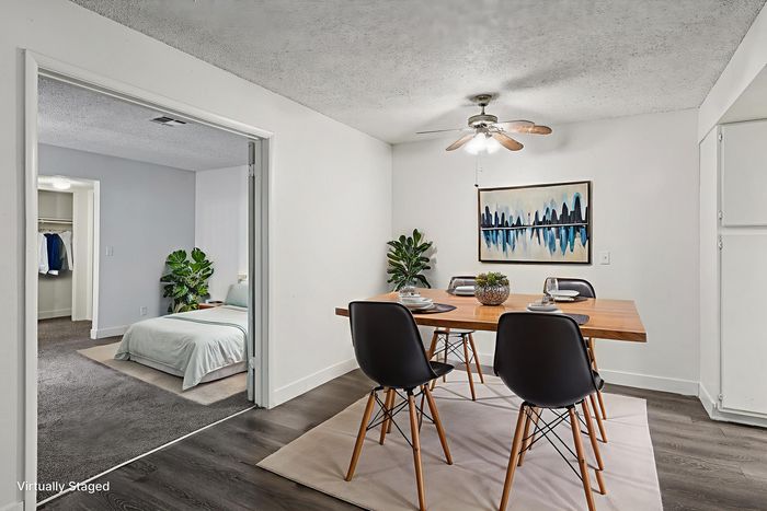 Elegantly staged apartment showcasing a stylish dining area with modern chairs and a wooden table, adjacent to a cozy bedroom with plush carpeting and fresh decor.
