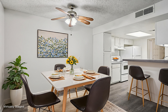Beautifully staged dining area with a modern table set for four, vibrant sunflower centerpiece, and adjacent open kitchen. Elegant decor with floral artwork and lush green plant enhancing the cozy atmosphere.