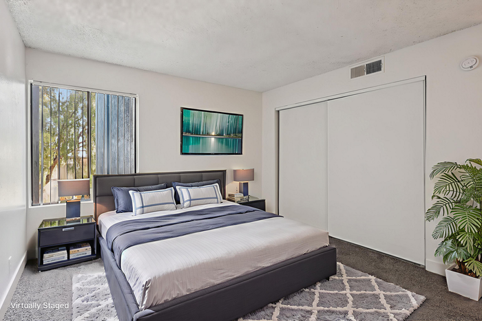 Elegant bedroom with modern decor, featuring a plush bed set against a serene artwork, natural light streaming through large windows, and a cozy ambiance enhanced by a stylish rug and vibrant green plant.