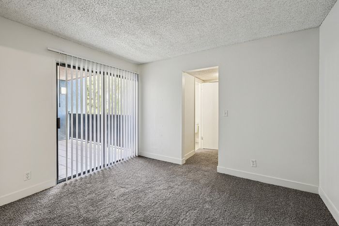 Bright and airy room with plush carpet, sliding glass door leading to a balcony, and a cozy corner leading to another room.