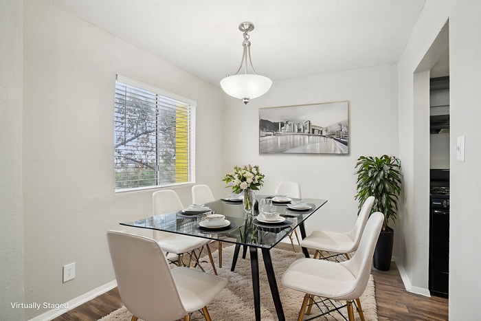 Elegant dining area with a modern glass table set for six, featuring chic white chairs, a stylish chandelier, and bright natural lighting from a large window.
