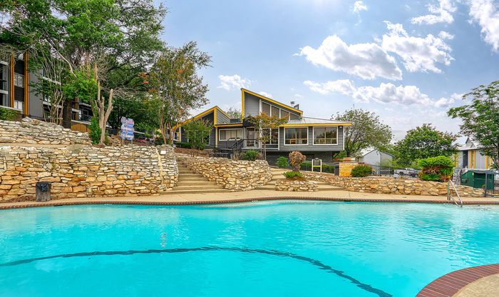 Charming apartment complex with modern architecture, surrounded by lush greenery and featuring a sparkling outdoor pool under a vibrant sky.