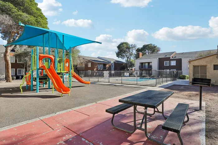 Charming apartment community featuring a vibrant playground with slides, a sparkling swimming pool, and cozy picnic area under blue skies.
