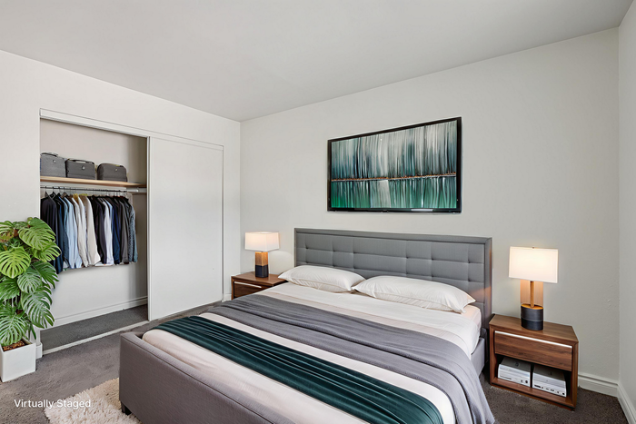 Elegantly staged bedroom featuring a plush gray headboard, modern bedside lamps, stylish artwork, and an organized open closet.