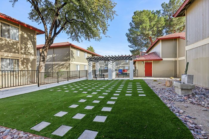 Charming apartment courtyard with lush green grass, sleek stone pathway, and a stylish pergola, framed by modern buildings with red-tiled roofs.