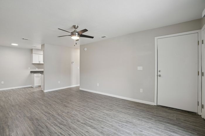 Chic modern living area with sleek hardwood flooring and a built-in ceiling fan, featuring an open layout leading to a stylish kitchen with contemporary white cabinetry.