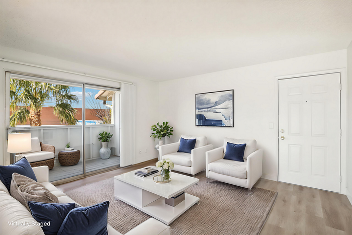 Chic living room with white modern furniture, plush rug, and large sliding glass doors opening to a balcony with palm tree views.
