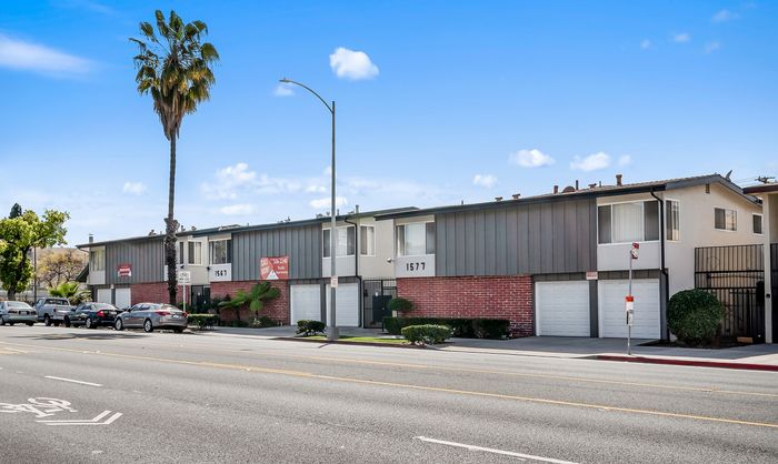 Charming two-story apartment complex with a modern exterior featuring red brick, green landscaping, and convenient parking under a clear blue sky.