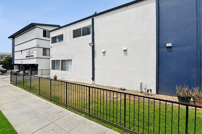 Modern apartment exterior with clean white and blue design, surrounded by a well-kept fence and green lawn, offering a stylish and inviting living space.