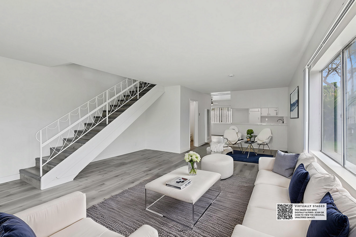 Modern living room with stylish staircase, cozy seating, and natural light, leading to a sleek kitchen area.