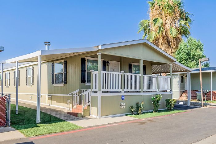 Charming mobile home with spacious porch, surrounded by lush greenery and a sunny, peaceful neighborhood setting.
