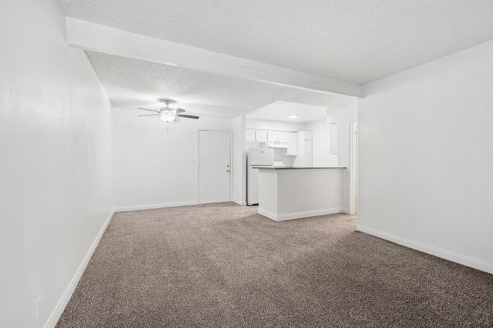 Bright living area with plush carpeting and open access to a modern kitchen, featuring a ceiling fan for added comfort.