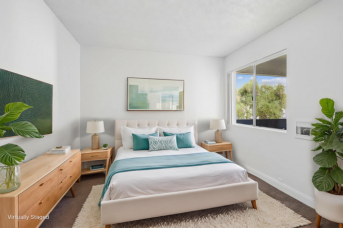Bright and airy virtually staged bedroom featuring a plush bed, modern wooden furniture, and a large window with lush greenery outside.