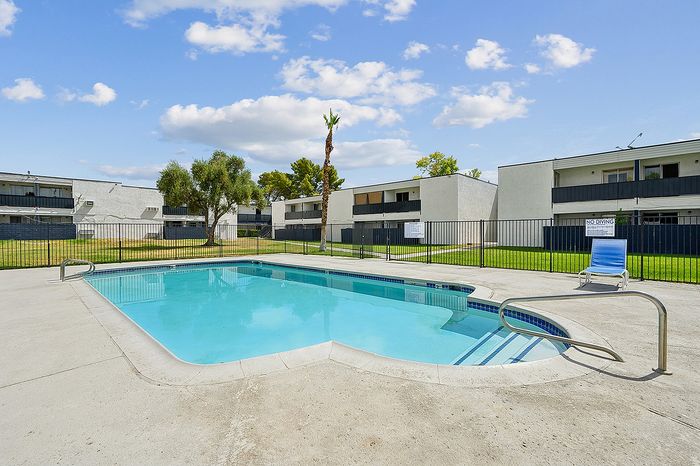Inviting outdoor pool area in a modern apartment complex, featuring clear blue water and a relaxing chair, set against a backdrop of lush greenery and contemporary buildings.