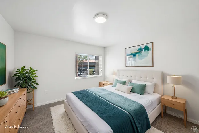 Bright and modern bedroom featuring elegant decor, a plush bed with teal accents, natural light from a window, and chic furnishings.