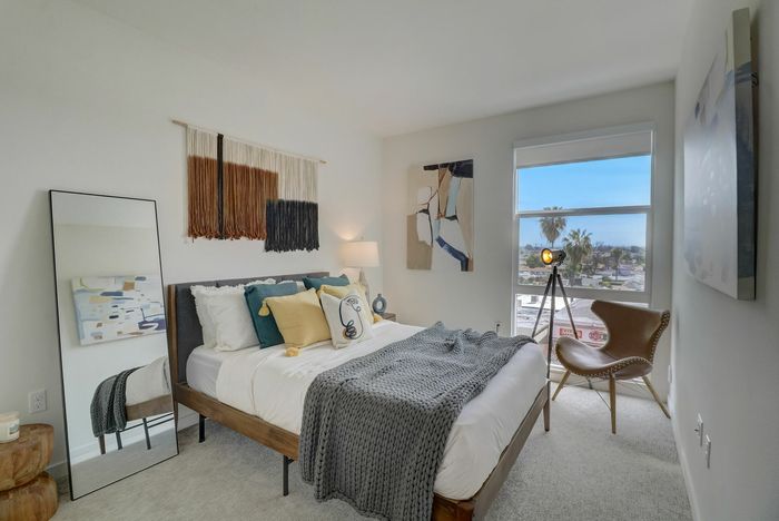 La Mesa, CA Apartments for Rent - Kelvin - Bedroom with Plush Carpeting, Large Bed, Full-Body Standing Mirror, Nightstand with Lamp, Corner Chair, and Large Window.