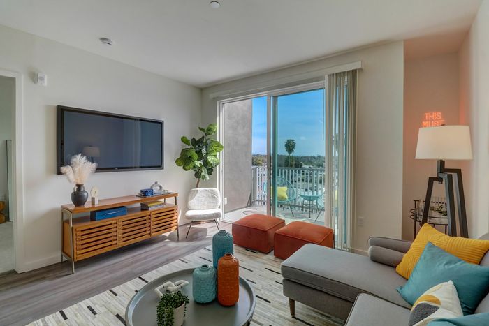 Apartments in La Mesa, CA - Kelvin - Living Room with Luxury Wood-Style Flooring, Area Rug, Sectional Couch, Round Coffee Table, Standing Lamp, Square Ottoman Stools, Chair, Wooden Cabinet, Wall-Mounted TV, Houseplant, and Sliding Glass Door with Balcony Access.