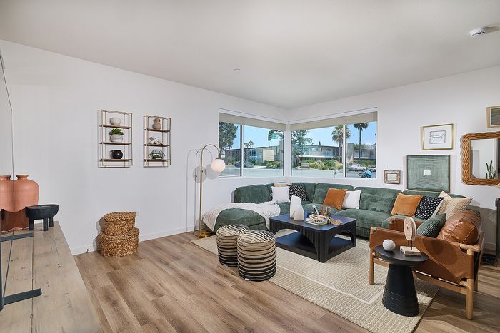Dog-Friendly Apartments in Serra Mesa, San Diego, CA - DEZI - Living Room with Durable Vinyl Plank Flooring, Corner Windows, Wall Art, Mirror, Sectional Couch, Coffee Table, Armchair, Side Table, Poufs, Floor Lamp, Storage Baskets, Wall Shelves, and Media Cabinet.
