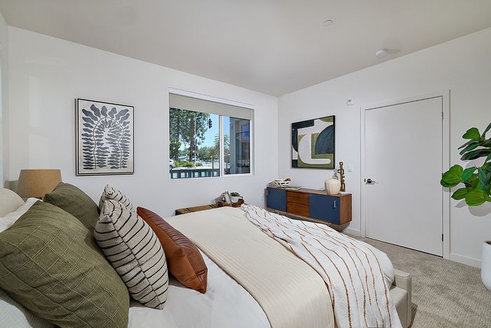 Apartments in Serra Mesa, San Diego, CA, With In-Unit Washer and Dryer - DEZI - Bedroom With Large Window, Neutral Bedding, Soft Carpet, Modern Wall Art, and Wood Dresser With Decor.