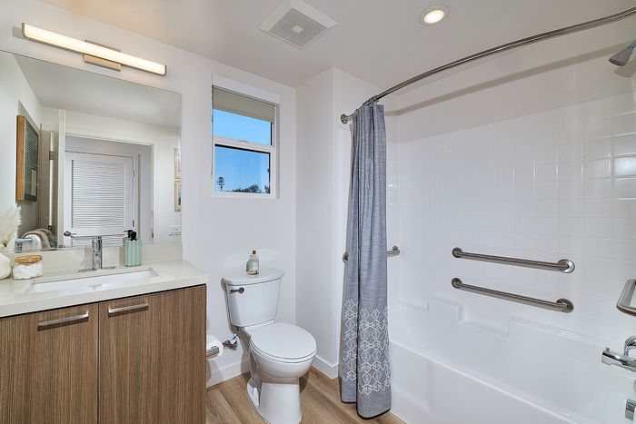 One-Bedroom Apartments in Serra Mesa, San Diego, CA - DEZI - Accessible Bathroom With Vanity Sink, Bright Mirror Lighting, Bathtub-Shower Combo With Grab Bars, and Window For Natural Light.