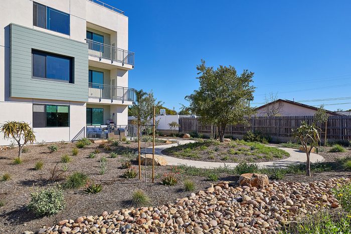 1-Bedroom Apartments in Serra Mesa, San Diego, CA - DEZI - Landscaped Courtyard With Winding Pathway, Native Plantings, Rock Garden, and Apartment Building Balconies Under Blue Sky.