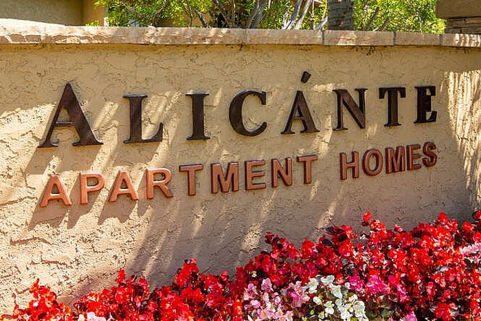 Close-up of the Elan Alicante Apartment Homes monument sign set in a stone wall with vibrant red and pink flowers in the foreground.