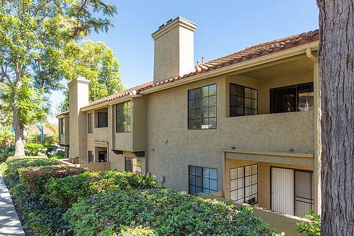 View of the two-story apartment buildings featuring Mediterranean-style architecture, tiled roofs, and private balconies nestled among mature trees.
