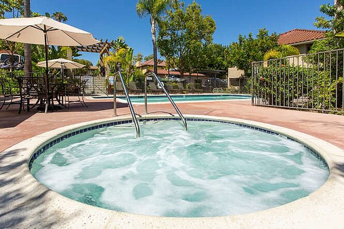 An outdoor circular spa with clear blue water and chrome handrails, situated next to a swimming pool with lounge seating and umbrellas.