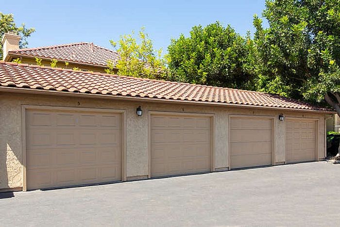 A row of private, attached garages with tan doors under a tiled roof, surrounded by lush green trees.