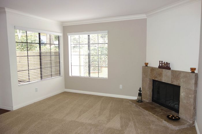 A bright apartment interior featuring neutral-toned carpeting, large windows, and a stone-faced fireplace.