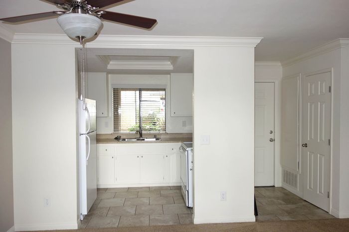 View from the living area looking into a kitchen with white appliances, wooden cabinetry, and durable flooring.