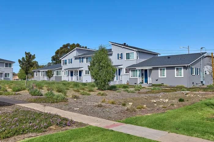 Village Apartments residences with yards, concrete pathways, and blue sky.