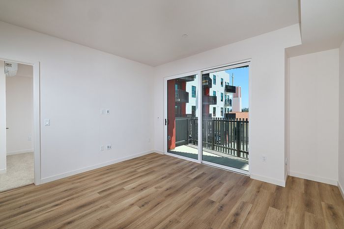Modern living room at textur in La Mesa with wood-style floors, sliding balcony door, and view into carpeted bedroom.