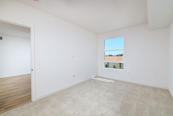 Carpeted bedroom in a textur La Mesa apartment with white walls and a window overlooking Spanish-style roofs.