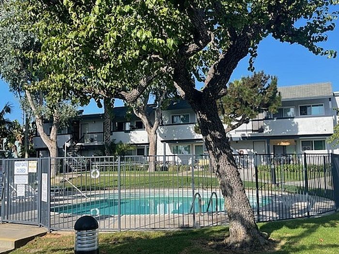 Fenced outdoor swimming pool shaded by trees in front of Delaware Pines apartment exterior.
