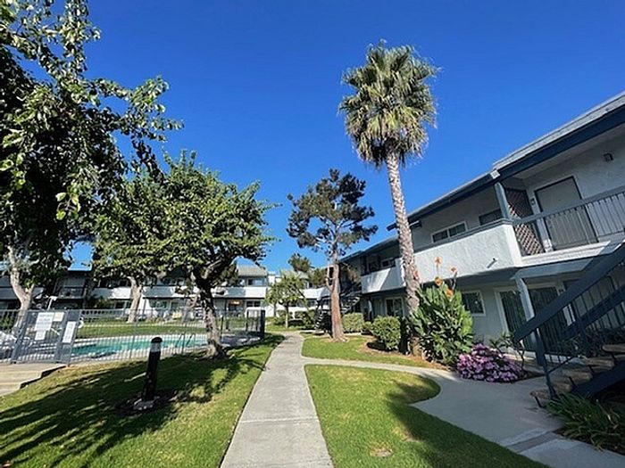 Fenced outdoor swimming pool shaded by trees in front of Delaware Pines apartment exterior.