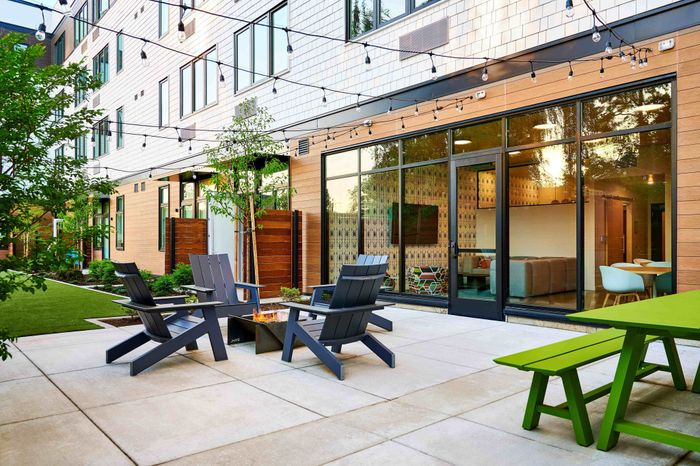 1 BR Apartments in Woodstock, Portland, OR - NOVUS - Outdoor Lounge Area With Fire Pit, Lounge Chairs, Picnic Table, String Lighting, And Well-Maintained Landscape.