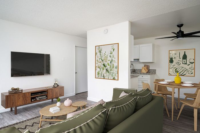 virtually staged living room with faux hardwood flooring and a green sofa with two coffee tables in front an entertainment console with a tv on the wall and a kitchen with a round dining table at boulder gardens