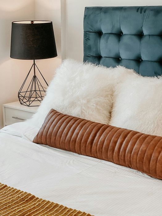 bed with fluffy accent pillows and leather tube pillow and a tufted blue suede headboard