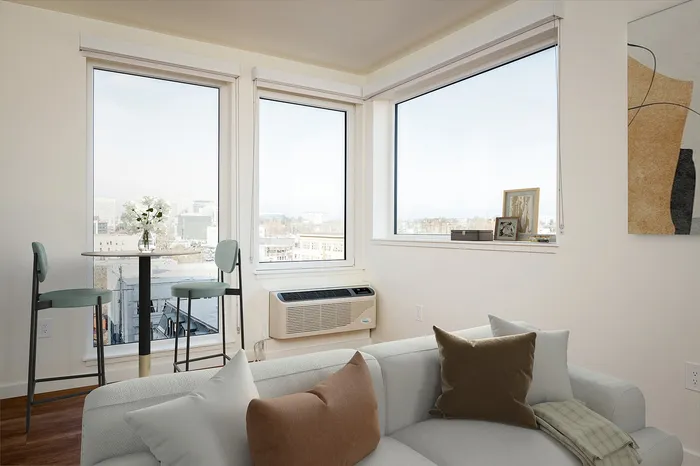 Pet-Friendly High Rise Apartments in Portland OR - Jute - Living Room with Expansive Windows, White Walls, a High-Top Dining Table with Stools, Wood-Style Flooring, and a Light Couchvirtual staged living room