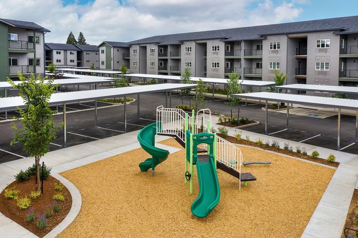 Pet-Friendly Apartments in Albany, OR - Creekside Meadows - Playground Featuring Green Slides and Safety Railings with Wood-Chip Ground, Surrounded by Carports and Building View.
