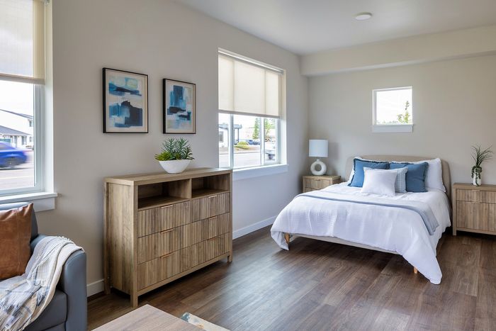 Albany, OR, Apartments - Creekside Meadows - Bedroom with Light-Colored Walls and Dark Wood Floors, Nightstands Flanking the Bed with a Lamp and Dresser, Potted Plants, Sofa, and Large Windows.