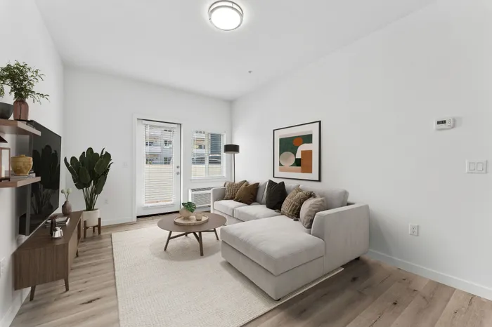 Newberg, OR, Apartments - Spring Meadow - Bright Living Room With Light Gray Sectional Sofa, Round Coffee Table, Wall Art, Wood Style Flooring, And Glass Door Leading To A Patio.