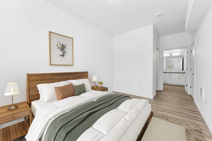1 Bedroom Apartments in Newberg, OR - Spring Meadow - Bright Bedroom With Wood Bed Frame, White Bedding, Matching Nightstands With Lamps, And An Open Hallway Leading To A Bathroom Vanity.