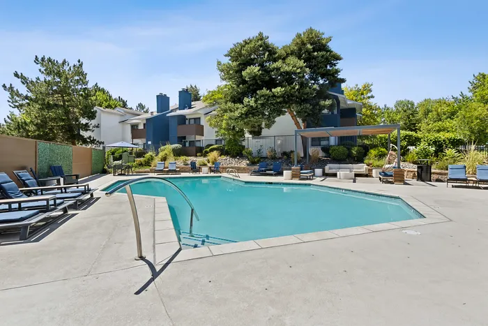 Dog-Friendly Apartments in South Reno, NV - Redfield Ridge - Resort-Style Outdoor Pool With Clear Blue Water, Poolside Lounge Chairs, Shaded Seating, Lush Trees, and Apartment Homes In The Background.