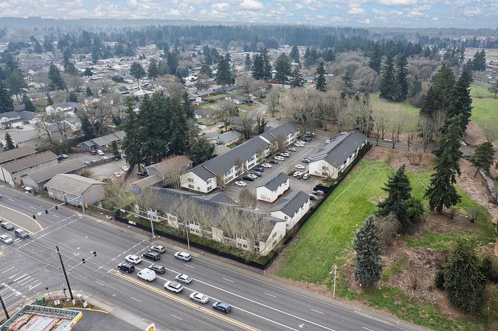 Aerial drone shot of property and surrounding area showing woodland with lots of trees, roads, gas station, and other local businesses as well as long horizon shot of woods and clouds.
