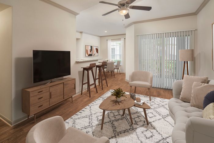 Elegant living room with modern decor featuring a plush sofa, chic coffee table, and a sleek TV stand. Natural light floods in through large windows with vertical blinds, highlighting the warm wooden flooring and stylish dining area with bar stools.